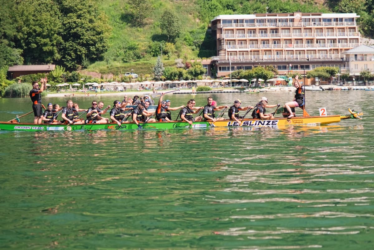 Una barca drago verde e gialla in gara sul lago di Levico, con gli edifici della riva alle spalle