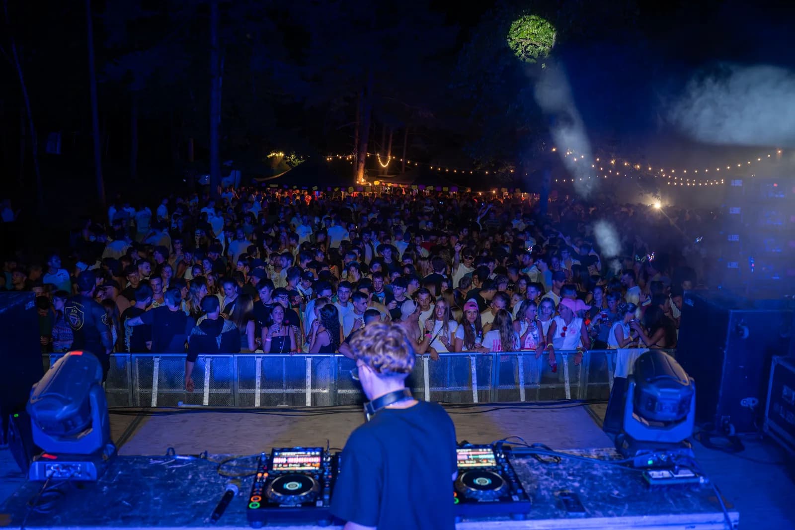 Un dj alla consolle di sera, ripreso da dietro, con il pubblico illuminato di blu
