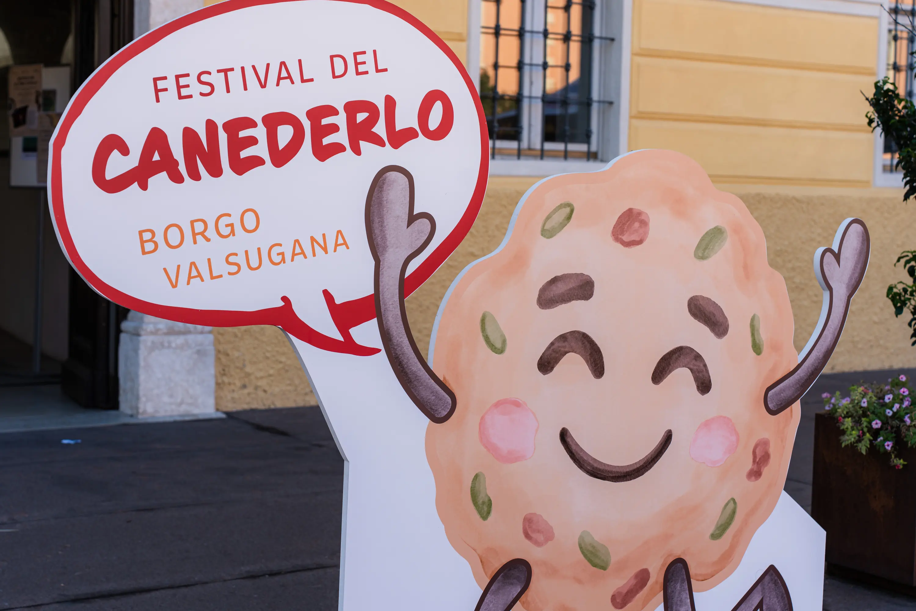 Il cartello del Festival del Canederlo con la mascotte, un canederlo sorridente a braccia alzate