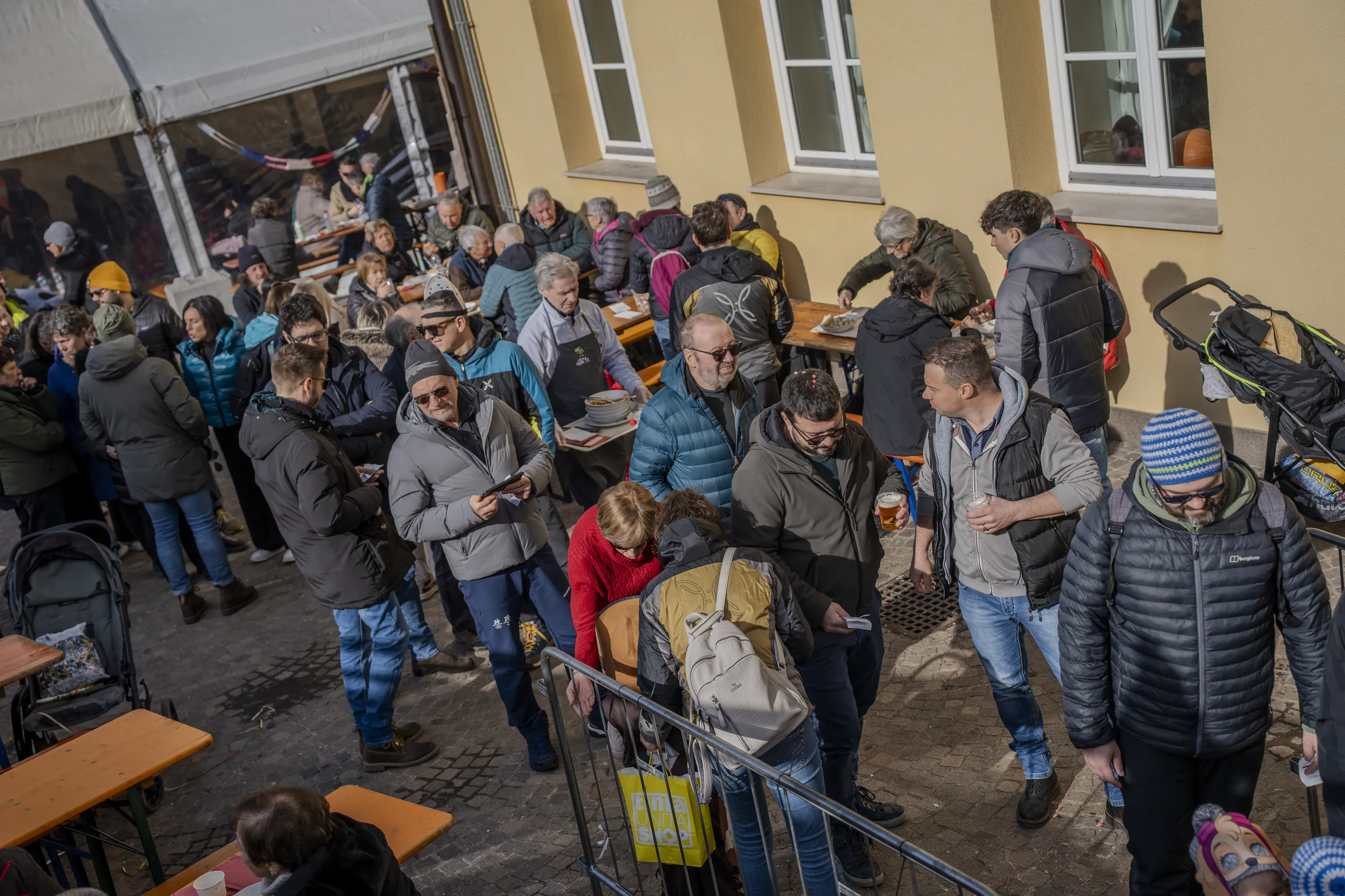 La via del paese con i tavoli e la gente in maschera, nella luce di febbraio