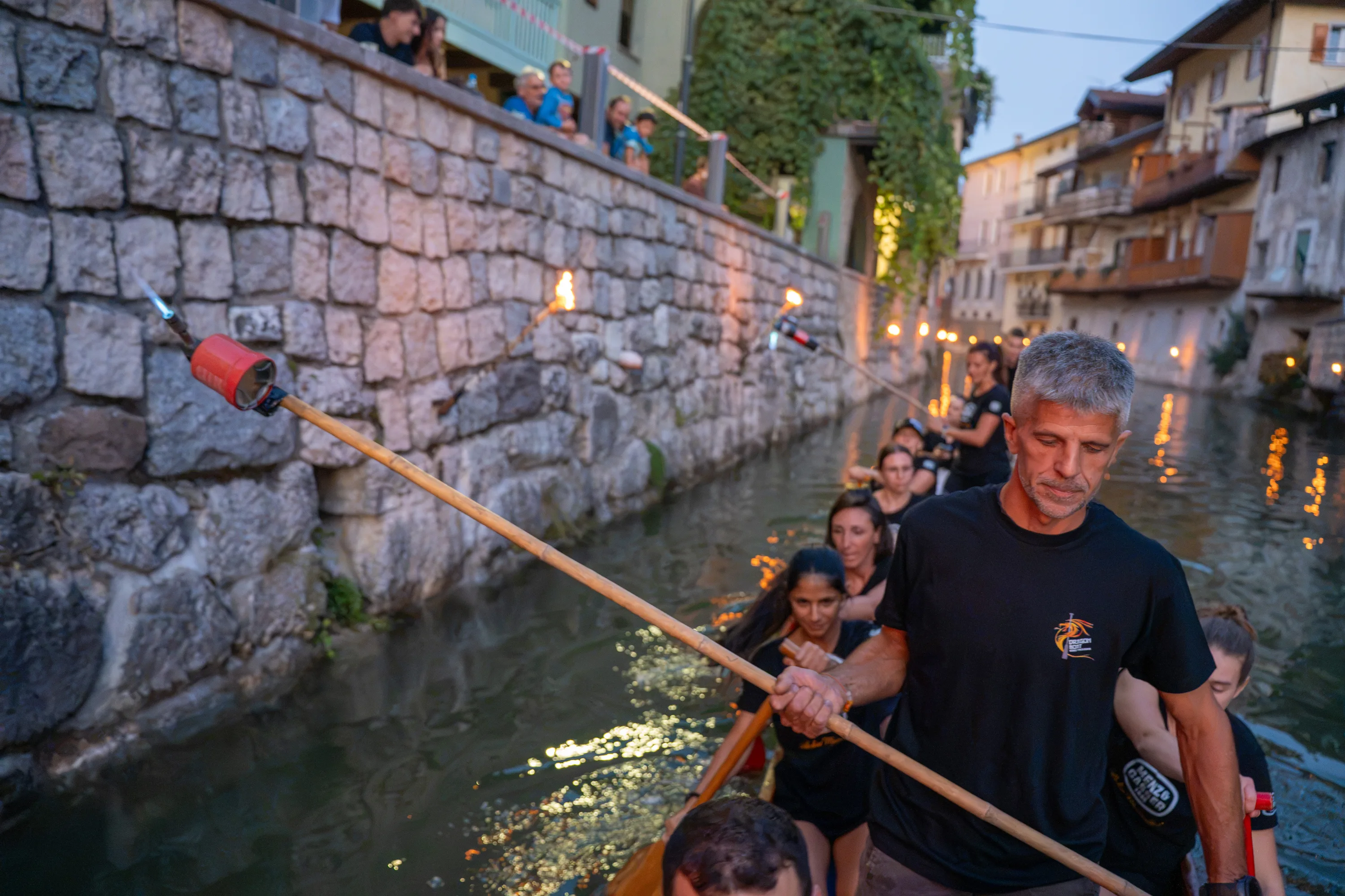 Dalla barca un uomo regge la lunga asta con il cannello per accendere le fiaccole sul muro, con l'equipaggio alle spalle