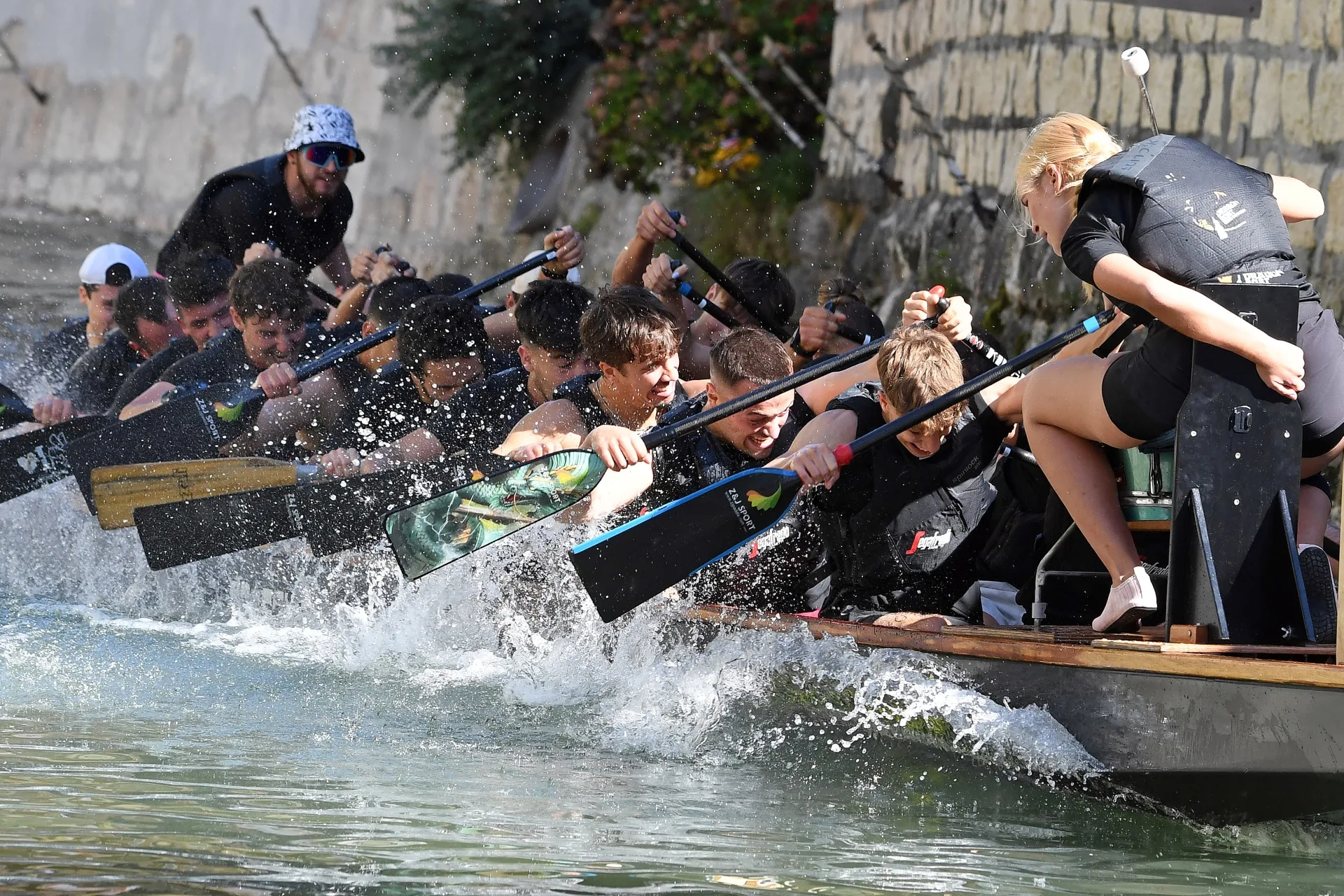 Un equipaggio affonda le pagaie nel Brenta lungo il muro di pietra, fra gli spruzzi, con la tamburina che si sporge verso la squadra