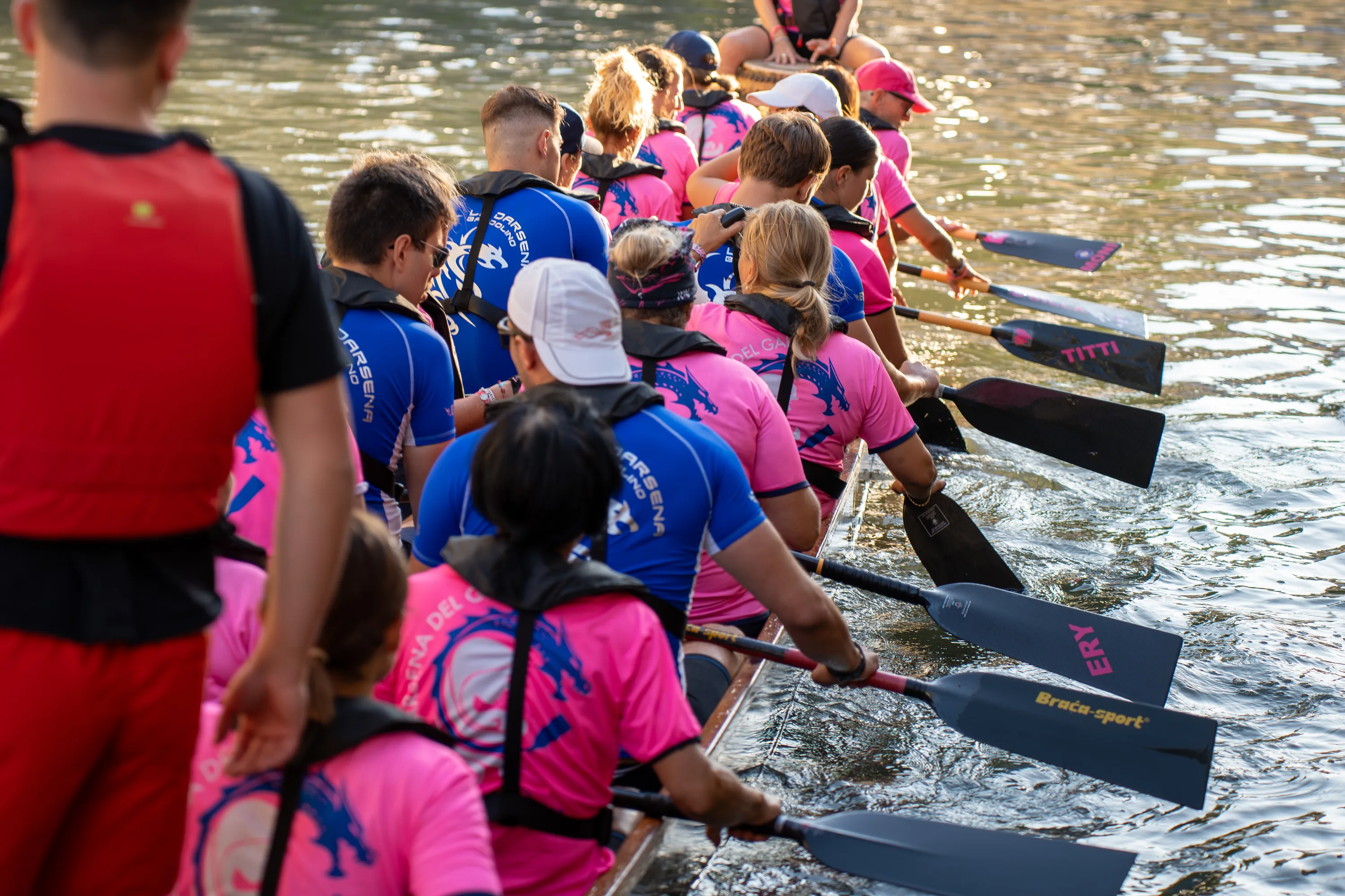 Le pagaie dell'equipaggio in rosa e blu entrano in acqua, viste alle spalle del timoniere